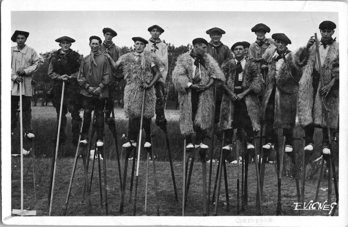 A black-and-white postcard photograph of more than a dozen men standing on tall stilts wearing berets, many in rustic sheepskin coats.